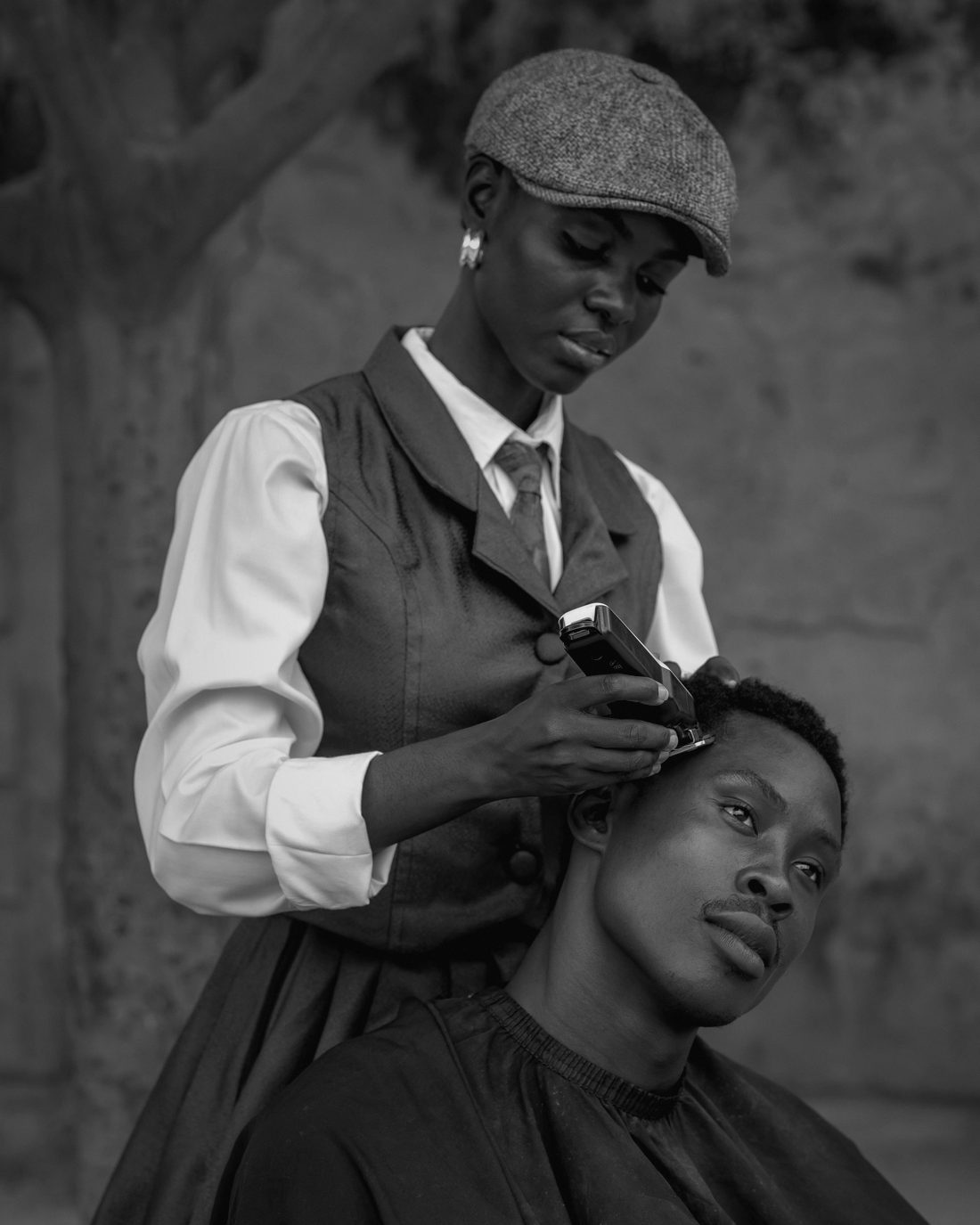 Black and white photo of a woman barber in a flat cap cutting a young man’s hair outdoors under a tree